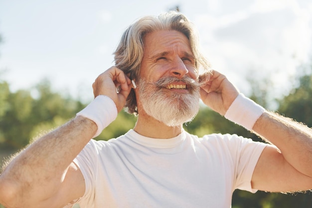 Escuchar música Senior hombre moderno y elegante al aire libre en el campo deportivo durante el día