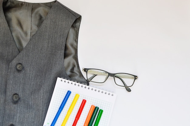 Escuela con chaleco, lápices, rotuladores y gafas sobre un fondo blanco.