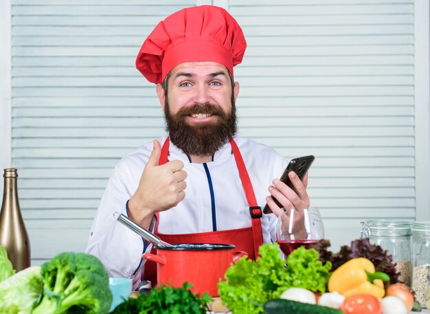 Escuela culinaria. Hipster con sombrero y delantal aprendiendo a cocinar en línea. Educación culinaria en línea. Concepto de elearning. Chef hombre buscando recetas de cocina para cocinar en Internet. Demostración culinaria del reloj del smartphone del cocinero.