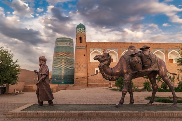 Escultura de camelo carregando mercadorias e homem de traje uzbeque em frente às muralhas da cidade de Khiva, Uzbequistão