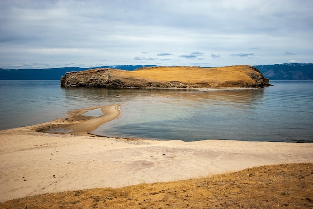 Escupir arena conduce a la isla en el lago Baikal. Detrás de las montañas, el cielo gris está encapotado. Hay arena en la orilla.