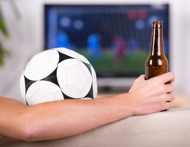 Esfera de futebol do close-up e mão com uma cerveja.