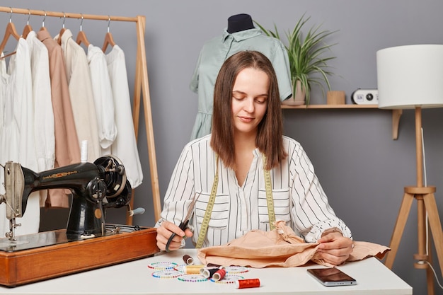 Esgoto de jovem concentrada concentrada sentada em seu local de trabalho no atelier perto da máquina de costura com uma tesoura nas mãos pensando em como alterar um vestido ou camisa velha
