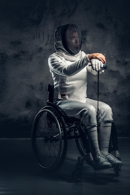 Esgrimista feminina em cadeira de rodas com máscara de segurança de um rosto, segurando florete em fundo cinza.
