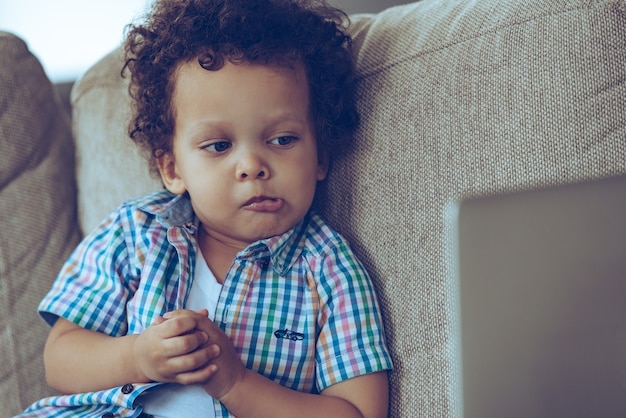 ¡Eso fue inesperado! Pequeño bebé africano mirando su computadora portátil y mirando confundido mientras está sentado en el sofá en casa