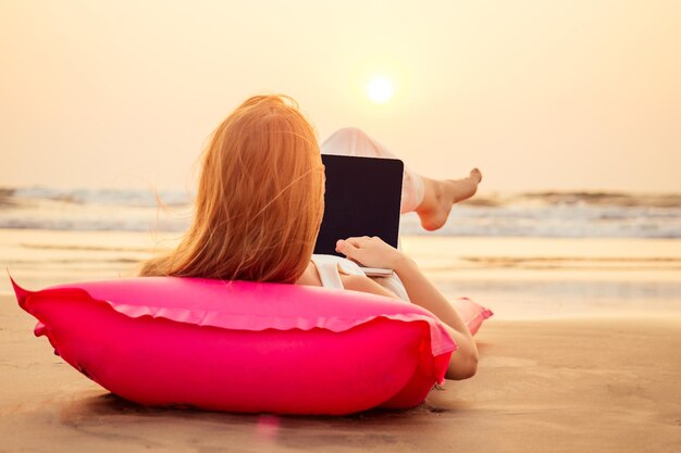 Espacio de copia Hermosa mujer joven que trabaja con una computadora portátil en la playa tropical Mujer viajera feliz usando una computadora portátil en vacaciones acostada en un colchón inflable rosa concepto de trabajo remoto