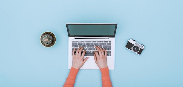 Espacio de trabajo azul claro minimalista con cámara digital portátil y cactus que una mujer está escribiendo en la vista superior del teclado