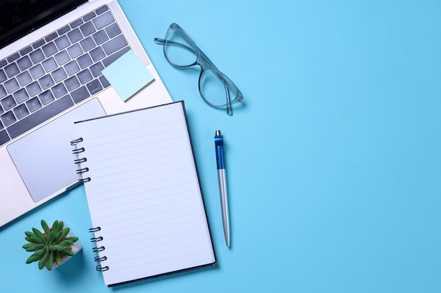 Espacio de trabajo de color azul de oficina con gafas para portátiles, notas adhesivas, mini bolígrafo suculento y página en blanco o