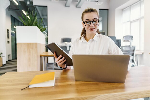 En un espacio de trabajo compartido, una joven dinámica equilibra hábilmente las tareas entre una computadora portátil y