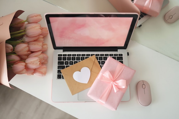 Espacio de trabajo de escritorio de estilo femenino con tulipanes rosas, computadora portátil y sobre. Vista superior de la pantalla del portátil. Caja de regalo rosa y ratón de ordenador rosa.