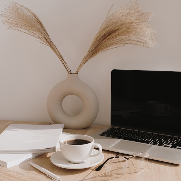 Espacio de trabajo de escritorio de oficina en casa femenina. Ordenador portátil de pantalla en blanco con espacio de copia. Taza de café, hierba de la pampa en elegante florero sobre mesa de madera beige.