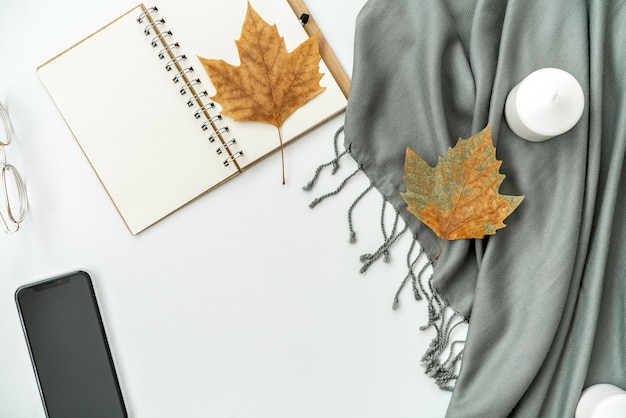 Espacio de trabajo de escritorio de oficina en casa con teléfono móvil con pantalla negra en blanco, cuaderno, hojas de arce, velas en blanco