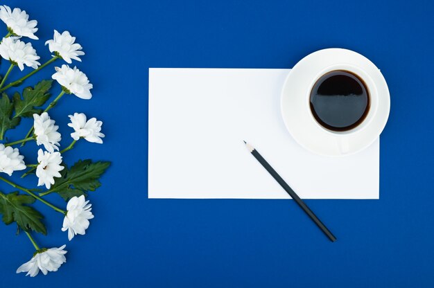 Espacio de trabajo femenino. Una taza de café y flores marco sobre fondo azul. Copia espacio