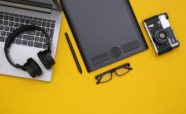 Espacio de trabajo de fotógrafo o retocador. Ordenador portátil, tableta gráfica, cámara y auriculares sobre fondo amarillo. Vista superior.