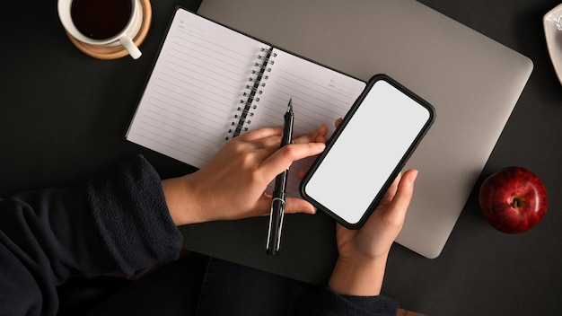 Espacio de trabajo hipster moderno con una pantalla blanca de teléfono inteligente en las manos de la mujer. vista superior