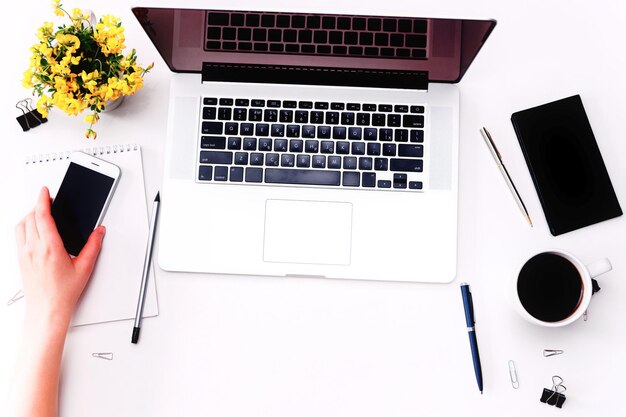 Espacio de trabajo con mano de mujer sosteniendo teléfono móvil, teclado portátil, café y flores amarillas en el fondo blanco. Plano, vista superior