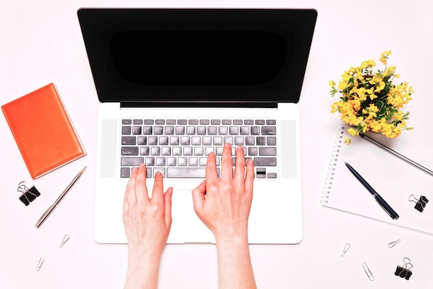 Espacio de trabajo con manos femeninas trabajando en teclado portátil, diario y flores amarillas en el fondo blanco. Plano, vista superior