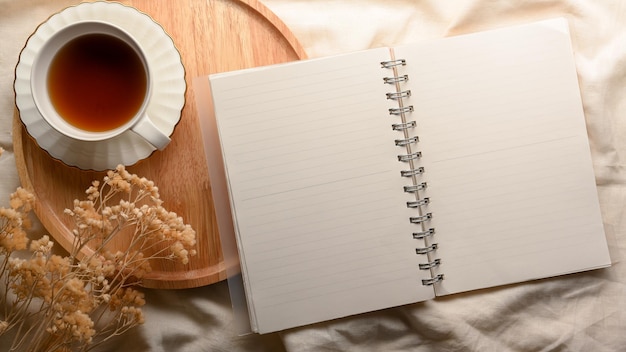 Espacio de trabajo mínimo con página de cuaderno vacía abierta, taza de té y flor seca sobre mantel blanco. plano