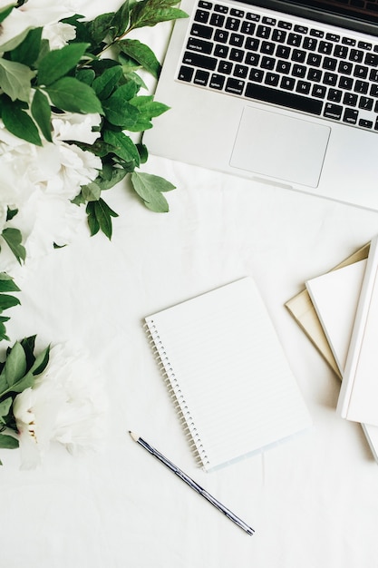 Espacio de trabajo de la oficina en casa con laptop, notebook, ramo de flores de peonías blancas sobre superficie blanca