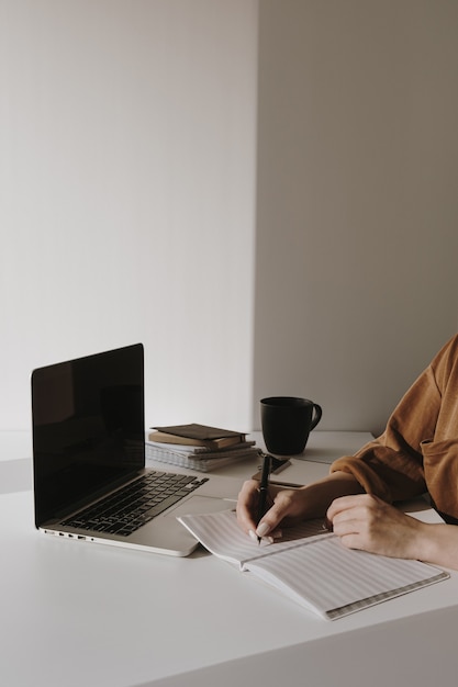 Espacio de trabajo de oficina en casa minimalista con computadora portátil, taza de café, portapapeles. Mujer escribe en un cuaderno de hojas de papel