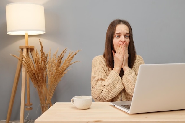 Foto espacio de trabajo profesional en línea asombrada mujer de cabello marrón con suéter beige trabajando en la computadora teniendo problemas con la conexión a internet mientras está sentada en la mesa en la oficina de casa