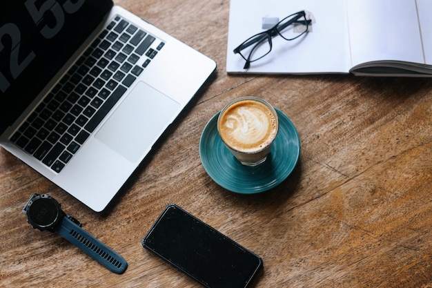 Espacio de trabajo con taza de café caliente, computadora portátil, anteojos y teléfono inteligente en la mesa de madera.