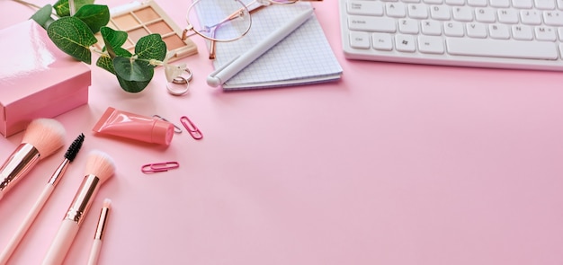 Espacio de trabajo con teclado, bloc de notas, gafas, bolígrafos, accesorios de belleza en concepto de superficie rosa