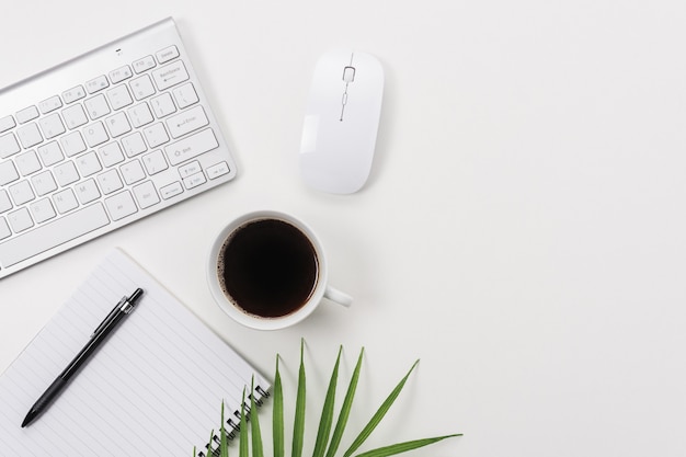 Espacio de trabajo con teclado de computadora, material de oficina, hoja verde y taza de café en blanco