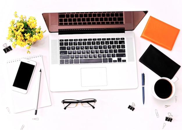 Espacio de trabajo y teclado portátil, teléfono móvil, café, vasos y flores amarillas en el fondo blanco. Plano, vista superior