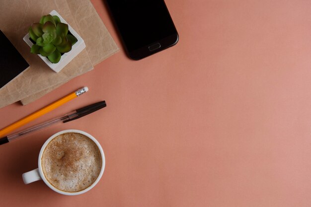 Espacio de trabajo con teléfono inteligente, material de oficina y taza de café sobre fondo coral.