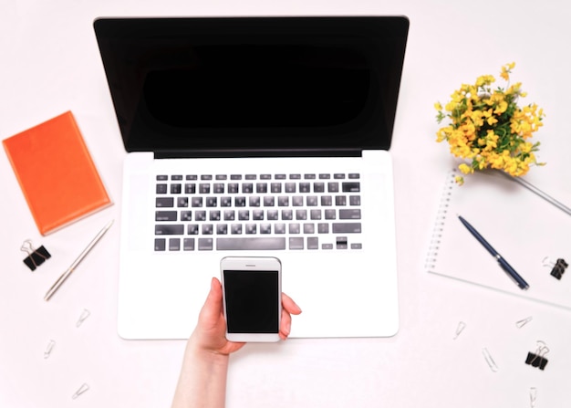 Espaço de trabalho com a mão de uma mulher segurando o celular e o teclado do laptop e flores amarelas no fundo branco. Postura plana, vista superior