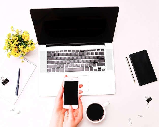 Espaço de trabalho com a mão de uma mulher segurando o celular no teclado do laptop, café e flores amarelas no fundo branco. postura plana, vista superior