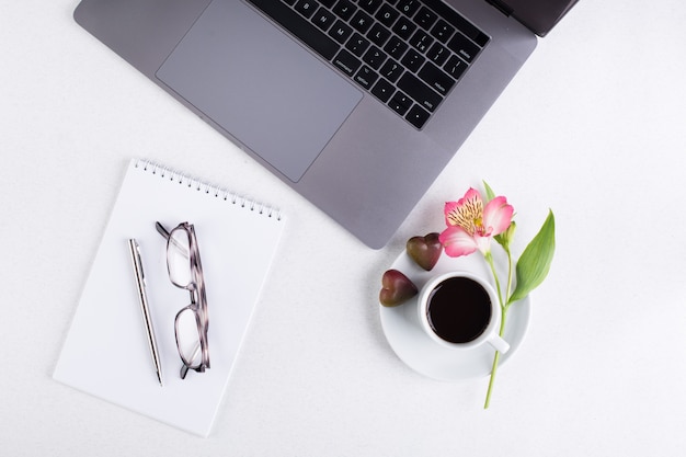 Espaço de trabalho com laptop, bloco de notas, tablet, óculos, xícara de café preto e uma flor de lírio peruano em um fundo branco