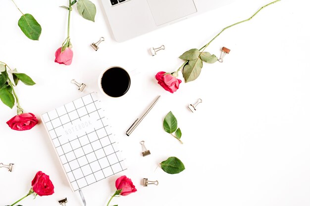 Espaço de trabalho com laptop, flores de rosas vermelhas, xícara de café, caderno e clipes na superfície branca