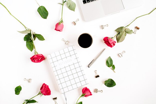 Espaço de trabalho com laptop, flores de rosas vermelhas, xícara de café, caderno e clipes na superfície branca