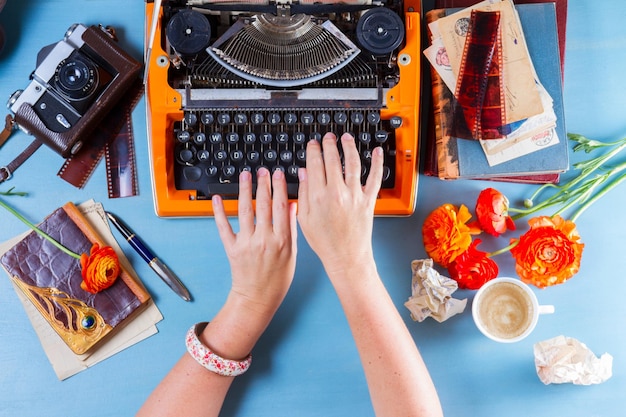 Espaço de trabalho com mãos de alguém digitando em máquina de escrever vintage laranja em fundo azul cena plana