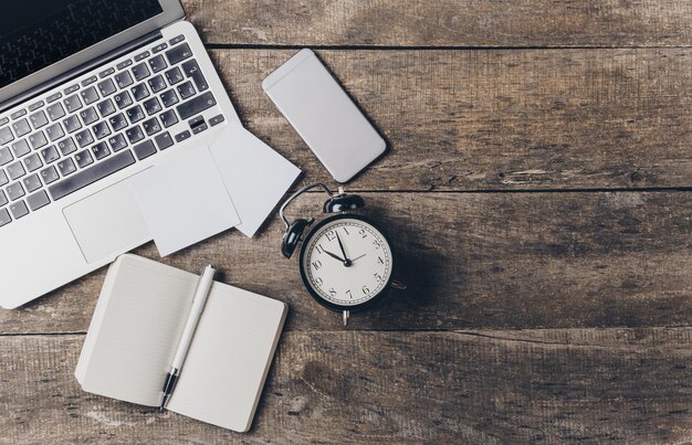 Espaço de trabalho com notebook de smartphone portátil e despertador em mesa de madeira durante as horas de luz do dia