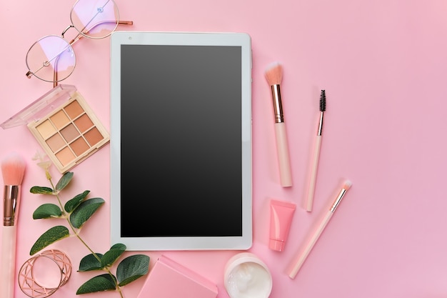 Foto espaço de trabalho com tablet branco, bloco de notas, óculos, canetas, acessórios de beleza, teclado, material de escritório, folha verde na superfície rosa