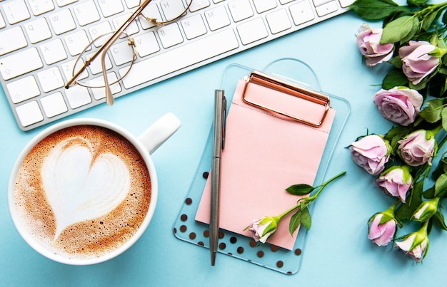 Espaço de trabalho com teclado, área de transferência, rosas em fundo azul pastel. mesa de escritório em casa. vista superior do fundo feminino. camada plana, vista superior.