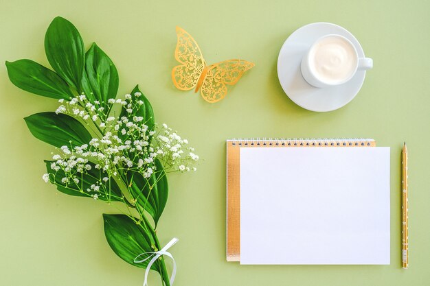 Espaço de trabalho com uma folha em branco de bloco de notas, lápis, folha verde, borboleta dourada decorativa e xícara de café sobre fundo verde.