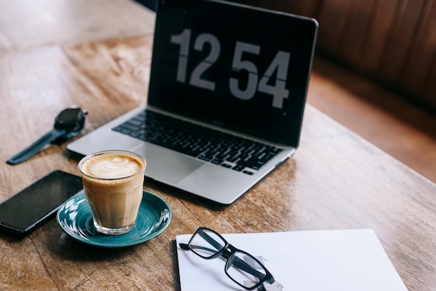 Espaço de trabalho com xícara de café quente, laptop, óculos e smartphone na mesa de madeira.