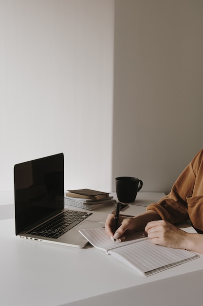 Foto espaço de trabalho de escritório em casa minimalista com laptop, xícara de café, área de transferência. mulher escrevendo em um caderno de papel
