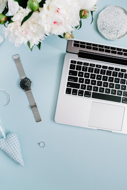 Espaço de trabalho feminino com laptop, buquê de flores de peônia branca, acessórios em azul