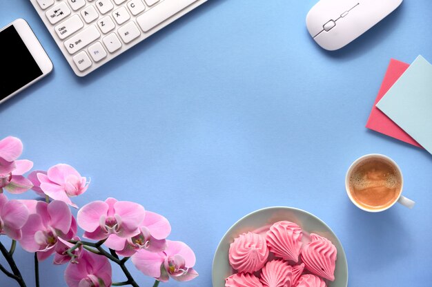 Espaço de trabalho feminino com orquídeas cor de rosa, teclado, telefone celular, prato de marshmallow e xícara de café