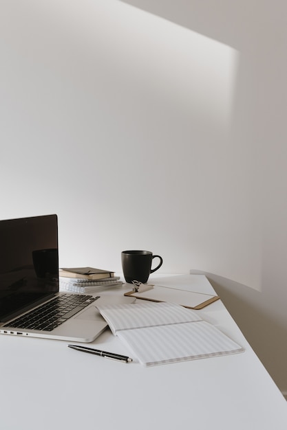 Espaço de trabalho minimalista da mesa do escritório em casa. Laptop na mesa com xícara de café, folha de papel, artigos de papelaria contra uma parede branca