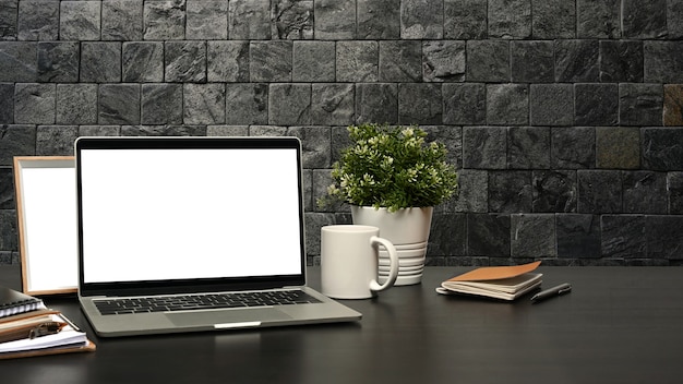 Espaço de trabalho moderno com computador portátil, xícara de café e planta em vaso na mesa preta com parede de tijolos.