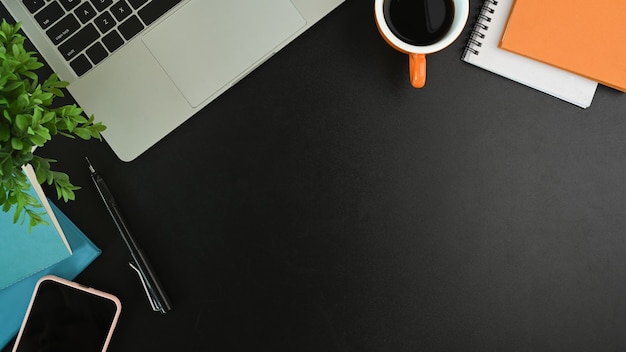 Espaço de trabalho moderno com laptop, telefone inteligente, xícara de café, caderno e planta na mesa preta.