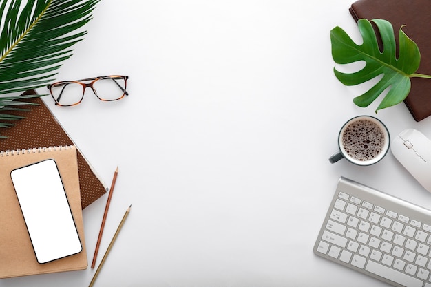 Espaço de trabalho moderno com teclado de computador e material de escritório. maquete de telefone móvel vazio na mesa branca com folhas de palmeira tropical. colocação plana do quadro.