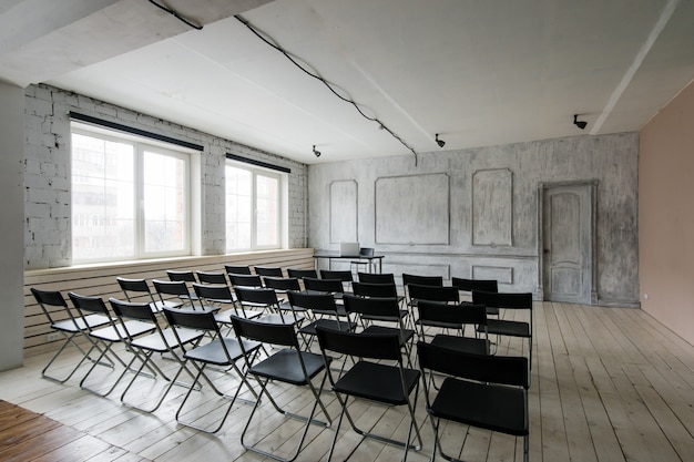 Espaço para palestras com muitas cadeiras escuras. As paredes são brancas, interior do sotão. À direita, há porta.