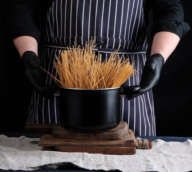 Espaguete longo amarelo cru em uma panela de metal preto e um chef em um avental listrado